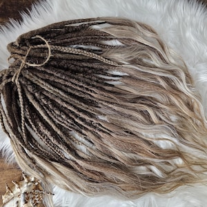 Może przedstawiać: Pęczek brązowych i blond dredów związanych sznurkiem. Dready są mieszanką ciemnobrązowych i jasnoblond, o falistej fakturze. Włosy leżą na białej, puszystej powierzchni.