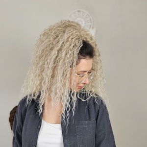 Op de afbeelding: Een persoon met krullend blond haar draagt een donkerblauw overhemd met knopen over een witte tanktop. Ze draagt een bril en kijkt naar beneden. Op de achtergrond is een witte ventilator te zien.