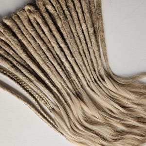 K&ouml;nnte beinhalten: Eine Ansammlung hellbrauner Dreadlocks, einige mit geflochtenen Enden, vor einem wei&szlig;en Hintergrund angeordnet. Die Dreadlocks haben ein nat&uuml;rliches, strukturiertes Aussehen mit verschiedenen Braun- und Blondt&ouml;nen. Das Bild zeigt die Textur und den Stil der Haarverl&auml;ngerungen.