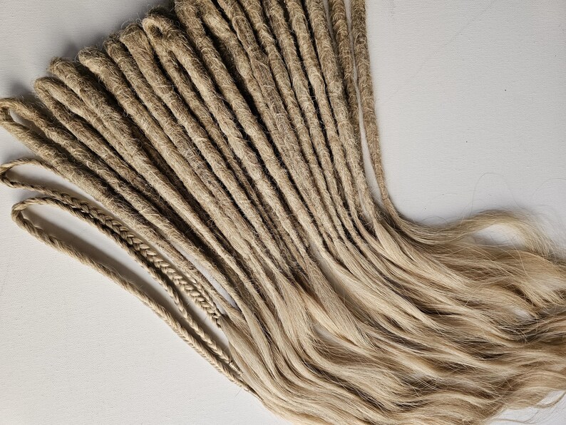 K&ouml;nnte beinhalten: Eine Ansammlung hellbrauner Dreadlocks, einige mit geflochtenen Enden, vor einem wei&szlig;en Hintergrund angeordnet. Die Dreadlocks haben ein nat&uuml;rliches, strukturiertes Aussehen mit verschiedenen Braun- und Blondt&ouml;nen. Das Bild zeigt die Textur und den Stil der Haarverl&auml;ngerungen.