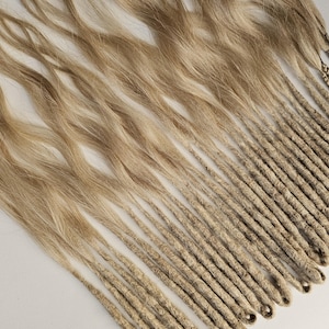 Peut inclure: Ensemble de dreadlocks blonds et brun clair, certains avec des cheveux lâches et ondulés en haut et d'autres étroitement enroulés. Les dreadlocks sont disposés en diagonale sur une surface blanche, avec quelques brins tressés visibles.