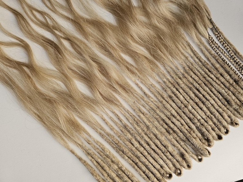 K&ouml;nnte beinhalten: Eine Sammlung von blonden und hellbraunen Dreadlocks, einige mit losem, welligem Haar oben und andere eng gewickelt. Die Dreadlocks sind diagonal &uuml;ber einer wei&szlig;en Oberfl&auml;che angeordnet, mit ein paar geflochtenen Str&auml;hnen.