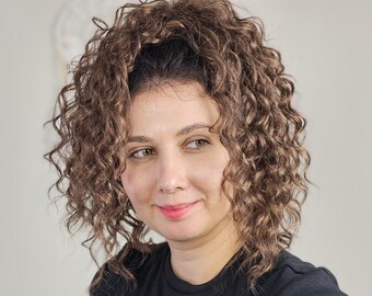 Könnte beinhalten: Eine schulterlange braune Perücke mit engen, voluminösen Locken und einem dunklen Wurzelansatz. Das Haar ist in einer runden Form gestylt und umrahmt das Gesicht mit strukturierten Ringellocken, die in einem Stufenschnitt fallen.