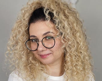 Könnte beinhalten: Eine Person mit voluminösem, schulterlangem, blondem Lockenhaar und dunklem Ansatz, die eine runde Brille mit schwarzem Rahmen und Metallbügeln trägt. Die Person ist mit einem schlichten weißen T-Shirt mit einem teilweisen schwarzen Grafikdruck bekleidet.
