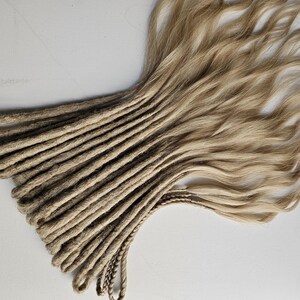 K&ouml;nnte beinhalten: Ein B&uuml;ndel blondierter Dreadlocks mit nat&uuml;rlicher, welliger Textur. Die Dreadlocks gehen von einer dunkleren, braunen Farbe an den Wurzeln zu einem helleren, goldblonden Farbton an den Enden &uuml;ber. Das Haar liegt auf einer wei&szlig;en Oberfl&auml;che.