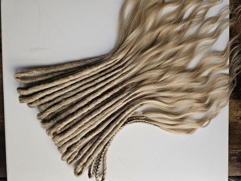 K&ouml;nnte beinhalten: Ein B&uuml;ndel blondierter Dreadlocks mit nat&uuml;rlicher, welliger Textur. Die Dreadlocks gehen von einer dunkleren, braunen Farbe an den Wurzeln zu einem helleren, goldblonden Farbton an den Enden &uuml;ber. Das Haar liegt auf einer wei&szlig;en Oberfl&auml;che.