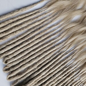 K&ouml;nnte beinhalten: Eine Sammlung hellbrauner Dreadlocks, diagonal auf einer wei&szlig;en Oberfl&auml;che angeordnet. Die Dreadlocks haben ein nat&uuml;rliches, strukturiertes Aussehen, wobei einige Str&auml;hnen wellig erscheinen. Die Farbe ist eine Mischung aus Hellbraun und Blond.
