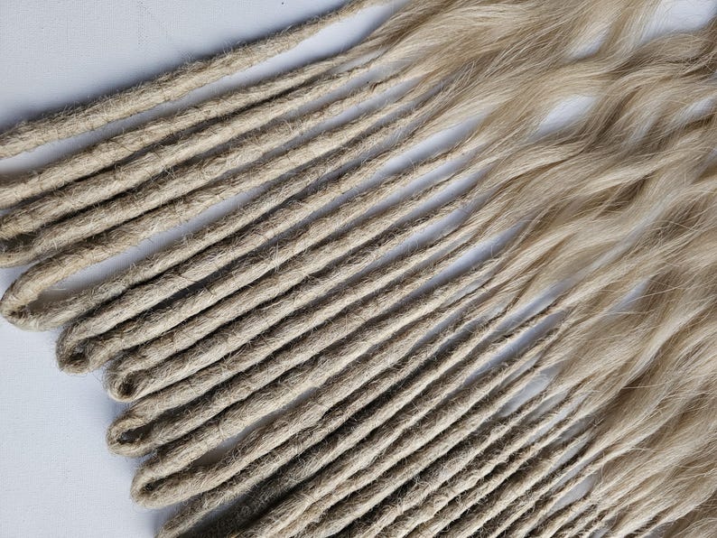 K&ouml;nnte beinhalten: Eine Sammlung hellbrauner Dreadlocks, diagonal auf einer wei&szlig;en Oberfl&auml;che angeordnet. Die Dreadlocks haben ein nat&uuml;rliches, strukturiertes Aussehen, wobei einige Str&auml;hnen wellig erscheinen. Die Farbe ist eine Mischung aus Hellbraun und Blond.