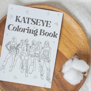 May include: A white coloring book titled "KATSEYE Coloring Book" featuring a group of outlined figures. The book is on a wooden plate with a cotton sprig, set against a neutral background.