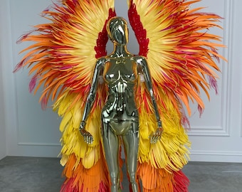 Può includere: Un manichino dorato mostra un vivace costume di piume. Il costume presenta grandi ali a strati nei toni del rosso, arancione e giallo, che ricordano una fenice. Le piume sono disposte per creare un effetto drammatico, perfetto per una festa in costume.