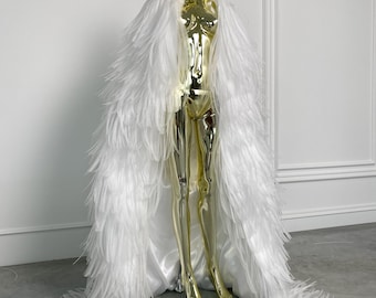 May include: A gold mannequin wearing a long, white feather cape. The cape drapes down to the floor, creating a dramatic effect. The mannequin is standing on a glass surface, and the background is a plain white wall.