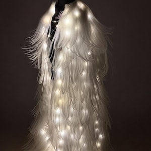 May include: A white, feathery garment illuminated with small, warm white lights. The costume is displayed on a black mannequin, creating a striking contrast. The design resembles angel wings or a flowing cape.