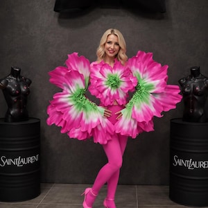 May include: A vibrant pink and green floral-inspired top featuring large, layered petal-like structures extending from the sleeves and chest, paired with matching pink leggings and pointed-toe heels. The ensemble is displayed in a setting with Saint Laurent branding.