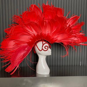 Op de afbeelding: Een levendige rode hoofdtooi met grote, gelaagde veren, gepresenteerd op een witte mannequinhoofd. De hoofdtooi heeft een decoratief ontwerp op het voorhoofd en rond het oog. De achtergrond is een donkergrijze, verticaal gestreepte muur.