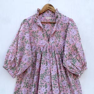 May include: A pink floral print dress with a V-neck and long, puffy sleeves. The dress features a gathered waist and a ruffled neckline. The floral pattern includes pink and purple flowers with green leaves.