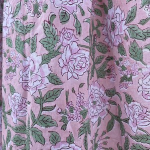 May include: Close-up of a fabric with a repeating floral pattern. Large, light pink roses with white centers and green leaves are printed on a light pink background. The fabric has a soft, textured appearance.