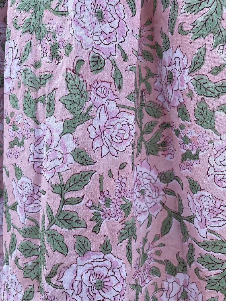 May include: Close-up of a fabric with a repeating floral pattern. Large, light pink roses with white centers and green leaves are printed on a light pink background. The fabric has a soft, textured appearance.