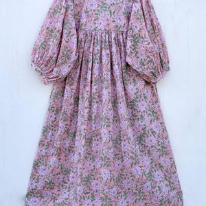 May include: A long, pink dress with a floral pattern. The dress features a high neckline, long sleeves, and a flowing skirt. The floral design includes purple and green accents, creating a soft, feminine look. The dress is hanging on a wooden hanger.