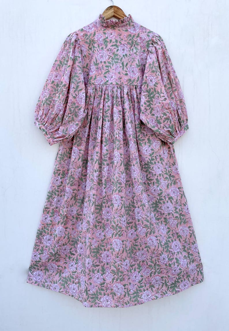 May include: A long, pink dress with a floral pattern. The dress features a high neckline, long sleeves, and a flowing skirt. The floral design includes purple and green accents, creating a soft, feminine look. The dress is hanging on a wooden hanger.