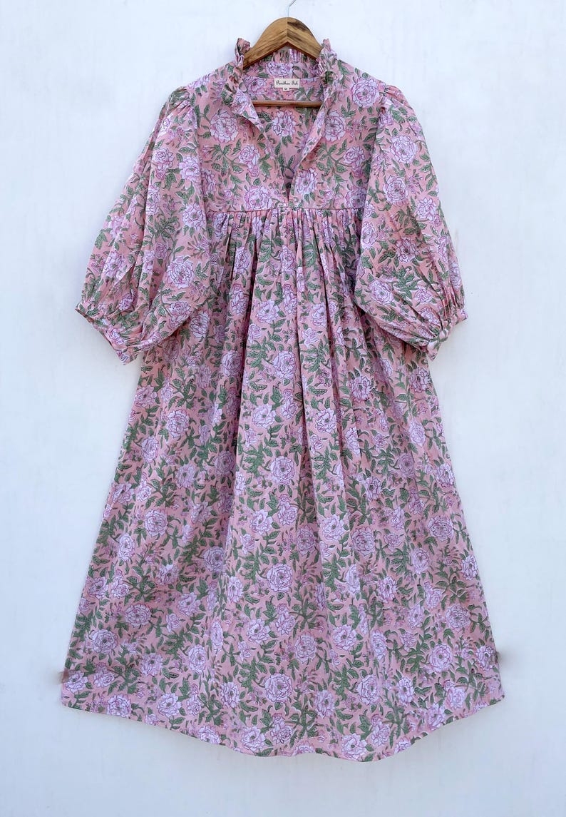 May include: A pink floral print dress with a V-neck and long, puffy sleeves. The dress features a gathered waist and a ruffled neckline. The floral pattern includes pink and purple flowers with green leaves.