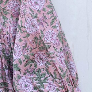 May include: A pink floral print dress with long sleeves. The fabric features a repeating pattern of large, light pink roses, green leaves, and smaller floral accents. The dress has a gathered neckline and a relaxed fit.