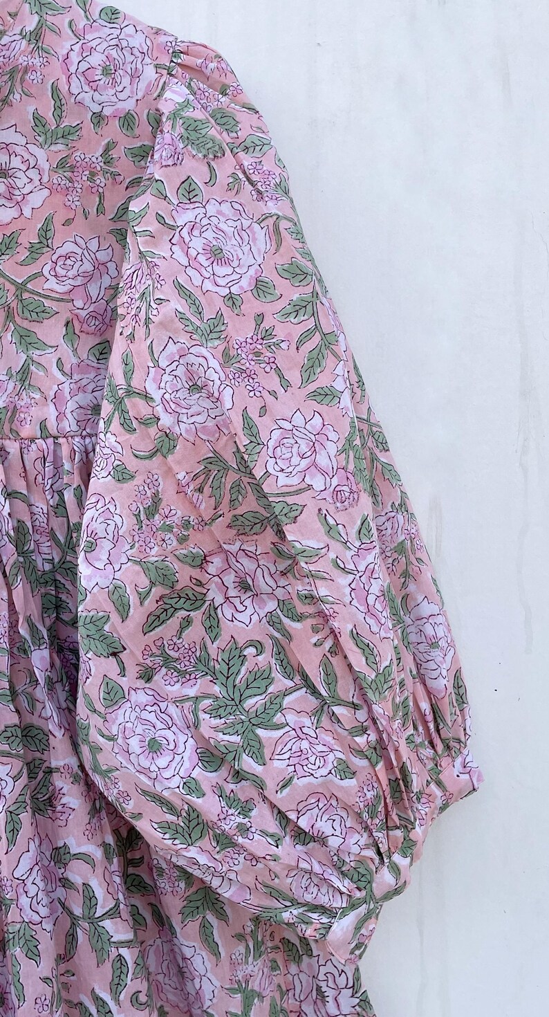May include: A pink floral print dress with long sleeves. The fabric features a repeating pattern of large, light pink roses, green leaves, and smaller floral accents. The dress has a gathered neckline and a relaxed fit.
