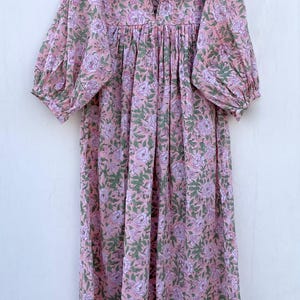 May include: A pink floral print dress with a ruffled neckline and long sleeves. The dress features a V-neck and a gathered waist. The fabric has a pattern of pink flowers and green leaves. The dress is hanging on a wooden hanger.