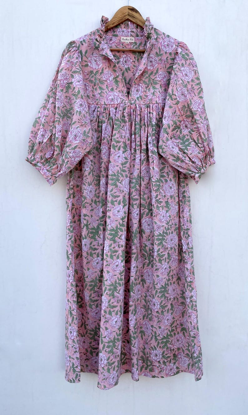 May include: A pink floral print dress with a ruffled neckline and long sleeves. The dress features a V-neck and a gathered waist. The fabric has a pattern of pink flowers and green leaves. The dress is hanging on a wooden hanger.