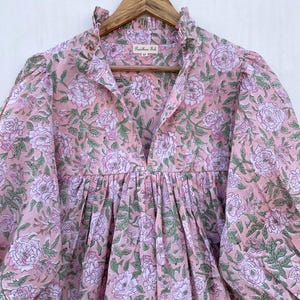 May include: A light pink floral print dress with a ruffled collar and long sleeves. The dress features a V-neckline and a gathered waist. The fabric is adorned with large, white and pink roses and green leaves. The dress is hanging on a wooden hanger.