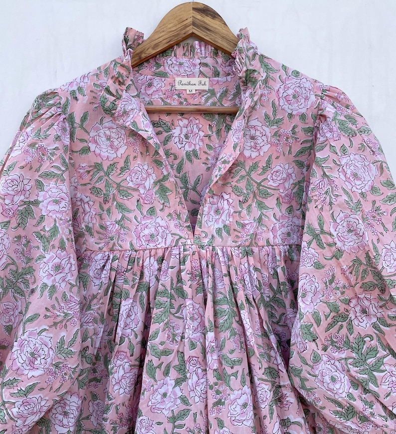 May include: A light pink floral print dress with a ruffled collar and long sleeves. The dress features a V-neckline and a gathered waist. The fabric is adorned with large, white and pink roses and green leaves. The dress is hanging on a wooden hanger.