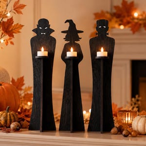 May include: Three black wooden candle holders shaped like silhouettes of a vampire, a witch, and a monster. Each figure features a small platform holding a white tea light candle. The figures have cut-out details for eyes and facial features.