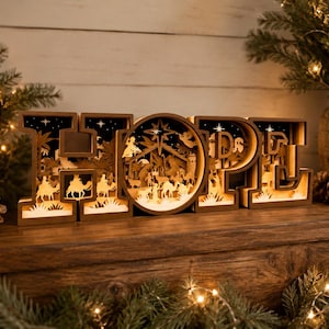 Pode incluir: Um letreiro decorativo de madeira com a palavra HOPE, apresentando silhuetas complexas cortadas a laser de uma cena de natividade dentro de cada letra. O design inclui camelos, figuras e uma estrela, iluminados por dentro com um brilho.