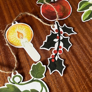 May include: A garland of paper cutouts featuring a red apple, holly with red berries, a lit candle, and green leaves. The cutouts are strung together with twine and are set against a brown wooden surface.