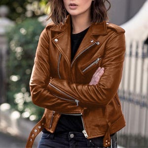 May include: A brown leather jacket with silver zips and a belt. The jacket features a classic biker style with a notched collar and multiple pockets. The jacket is a warm brown colour and appears to be made of soft leather.