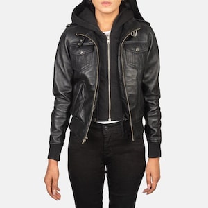 May include: Black leather jacket with a zip-up front, multiple pockets, and a hooded sweatshirt underneath. The jacket has a bomber-style fit with ribbed cuffs and a waistband. The model is wearing black trousers.