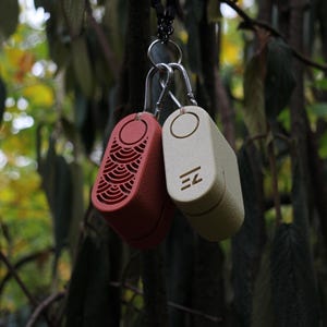 May include: Two small, rectangular, keychains hang from a metal ring. One is red with a decorative pattern, the other is off-white with the letters "EZ". The keychains are suspended against a blurred background of green leaves.