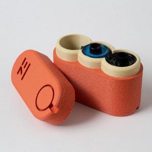 May include: An orange, textured container with three cylindrical openings, each with a different colored interior. A separate orange lid with a circular cutout and etched design is next to the container. The design appears to be a film canister.