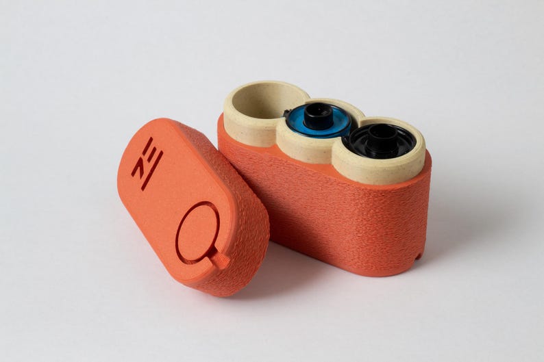 May include: An orange, textured container with three cylindrical openings, each with a different colored interior. A separate orange lid with a circular cutout and etched design is next to the container. The design appears to be a film canister.