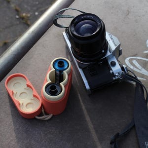 May include: A silver and black Canon camera with a black lens and strap sits next to a red and cream-colored film container. The container is open, revealing film rolls. The scene is set on a concrete surface.
