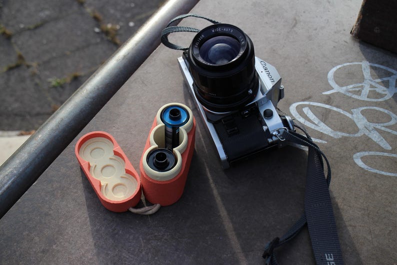 May include: A silver and black Canon camera with a black lens and strap sits next to a red and cream-colored film container. The container is open, revealing film rolls. The scene is set on a concrete surface.