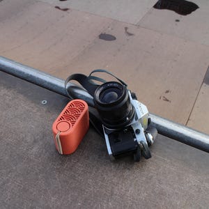 May include: A silver Canon camera with a black lens and strap rests next to an orange rectangular speaker. The speaker has a circular button and a patterned design on the front. The items are on a brown surface.