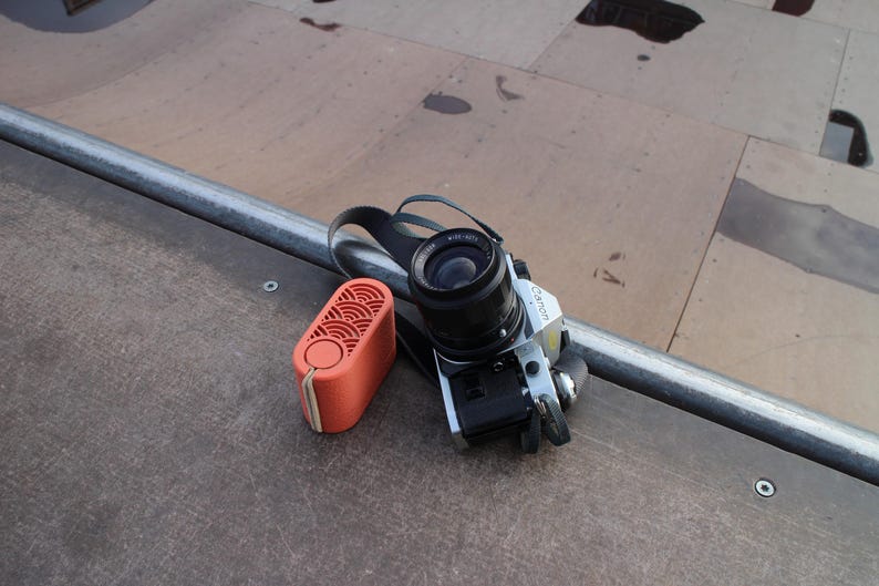 May include: A silver Canon camera with a black lens and strap rests next to an orange rectangular speaker. The speaker has a circular button and a patterned design on the front. The items are on a brown surface.