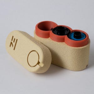 May include: A beige, rectangular container with three circular openings, each with a different colored interior: black, blue, and empty. The lid, also beige, is detached and features a circular handle and a stylized symbol.