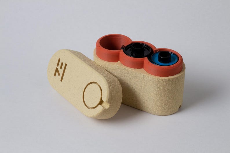 May include: A beige, rectangular container with three circular openings, each with a different colored interior: black, blue, and empty. The lid, also beige, is detached and features a circular handle and a stylized symbol.