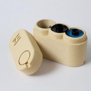 May include: Beige film canister with a matching lid. The canister has a textured surface and holds two film rolls with black and blue centers. The lid features a circular indentation and the letters "ZE".