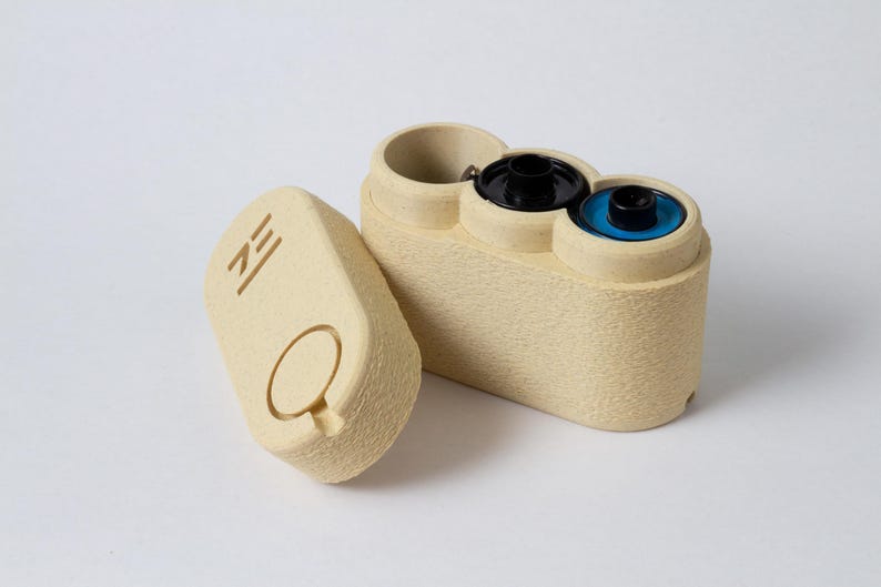 May include: Beige film canister with a matching lid. The canister has a textured surface and holds two film rolls with black and blue centers. The lid features a circular indentation and the letters "ZE".