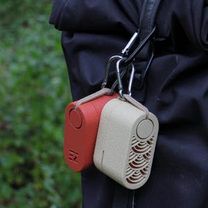 May include: Two small, rectangular, portable speakers are attached to a black strap with silver carabiner clips. One speaker is red, the other is beige. The red speaker has the letters "EZ" printed on it. The beige speaker has a wave pattern.