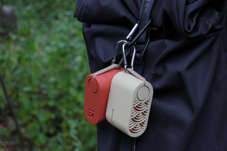 May include: Two small, rectangular, portable speakers are attached to a black strap with silver carabiner clips. One speaker is red, the other is beige. The red speaker has the letters "EZ" printed on it. The beige speaker has a wave pattern.