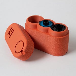 May include: An orange 3D-printed film canister with a textured surface. The canister is open, revealing two film rolls and a third empty compartment. The lid is separate, with a circular design and the letters "ZE" printed on it.