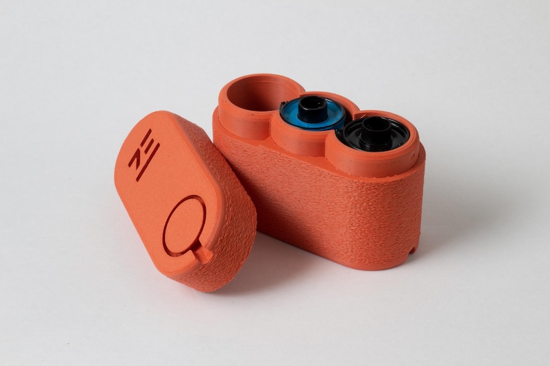 May include: An orange 3D-printed film canister with a textured surface. The canister is open, revealing two film rolls and a third empty compartment. The lid is separate, with a circular design and the letters "ZE" printed on it.